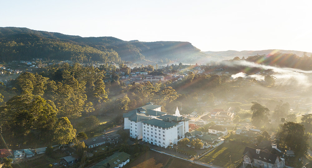 這張圖片展示了亞拉利亞綠城酒店（Araliya Green City Hotel）的全景。酒店位於斯里蘭卡高山地區的努瓦拉艾利耶（Nuwara Eliya），被茂密的森林和寧靜的山谷環繞。清晨的陽光透過雲霧照射在酒店建築上，營造出一個寧靜而優雅的氛圍，完美呈現了這個風景如畫的度假勝地的迷人景觀。