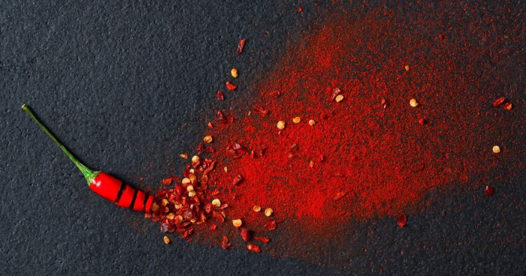這張圖片展示了一根紅辣椒及灑出的辣椒粉，深紅色的辣椒粉鋪展在黑色表面上，展現出濃烈的辛辣感。