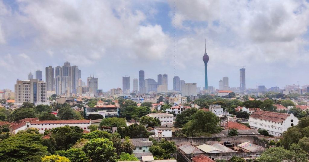 一個晴朗的天空下，可倫坡市中心的高樓大廈與特色建築映照在蔥鬱的綠樹背景中，展現了斯里蘭卡首都的現代都市氛圍。