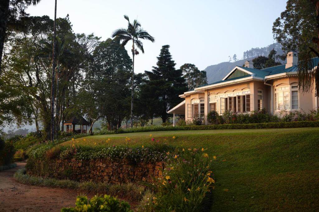 斯里蘭卡的殖民風格莊園位於山區，周圍綠意盎然。白色的建築物外觀古典，充滿歷史氣息，花園中種滿色彩繽紛的花卉，背景為蔥鬱的樹林與藍天，營造出寧靜的度假氛圍。
