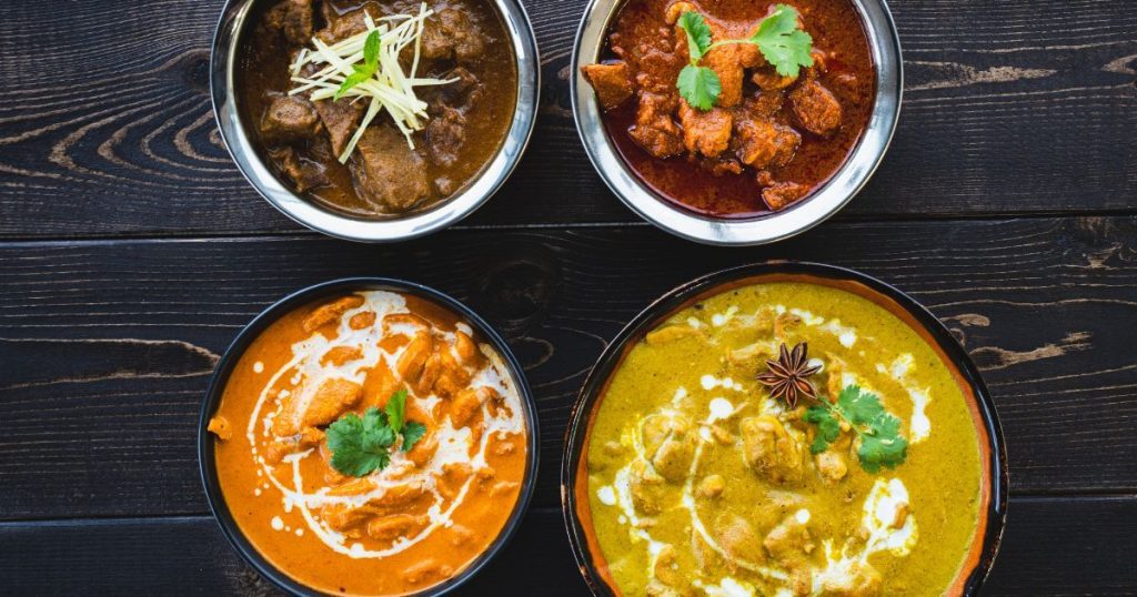 四碗色彩鮮豔的咖哩,從紅色到橙色再到綠色,餐盤上裝飾著香草,擺放在深色木質餐桌上。