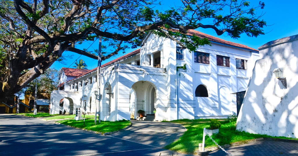 圖片展示位於斯里蘭卡加耳（Galle）的白色殖民建築——法院大樓。建築結構古典優雅，屋頂覆蓋紅瓦，周圍有翠綠的草坪和高大的樹木，陽光穿透枝葉形成美麗的光影效果，反映斯里蘭卡殖民時期的歷史建築風格與當地的熱帶自然風情。