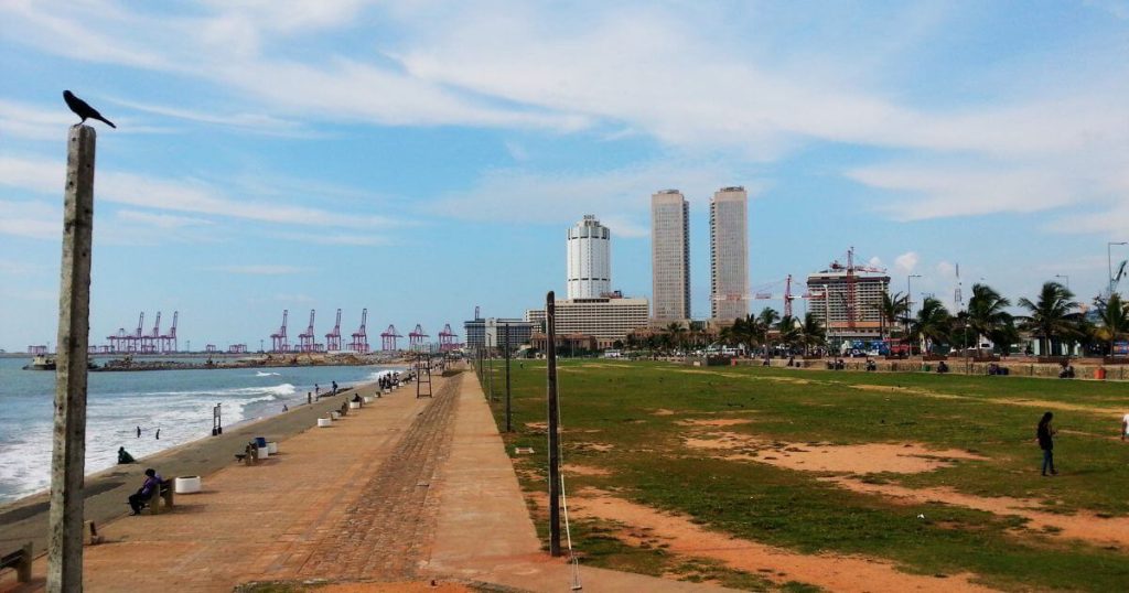 圖片展示了斯里蘭卡可倫坡著名的Galle Face Green海濱公園，公園綠地與遠處的高樓和港口構成了寬闊的景觀，遊客可以在海邊漫步，享受陽光與海風的舒適氛圍。