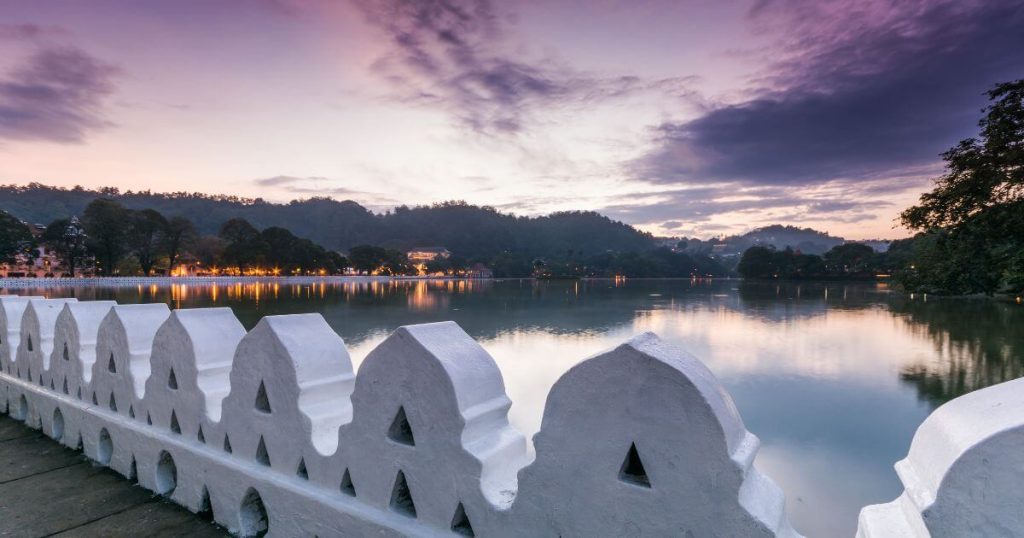 該圖片展示了斯里蘭卡康提湖的傍晚美景。湖面倒映著落日的餘暉,周圍的山脈與樹木剪影映襯在水中,讓整個畫面充滿寧靜氛圍。前景中的白色圍欄帶來了傳統建築風格的特色,展現了康提的自然與人文融合之美。