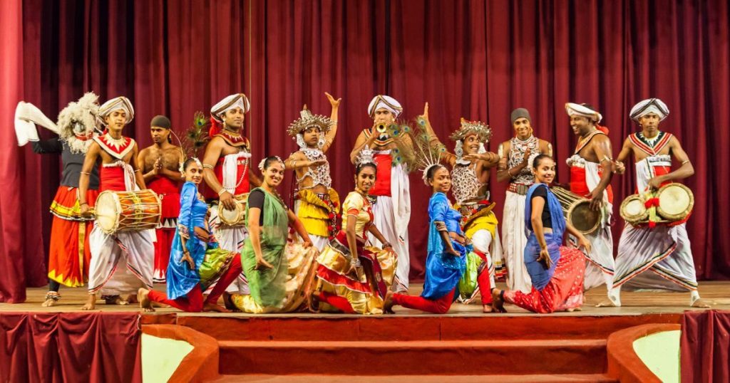 這張圖片展示了一群穿著色彩鮮豔傳統服飾的舞者,進行斯里蘭卡康提舞蹈表演。舞者們的動作充滿力量和優雅,身穿紅、藍、綠等色彩的服裝,搭配各種傳統樂器,包括大鼓和小鼓,背景是一道深紅色的舞台布幕。