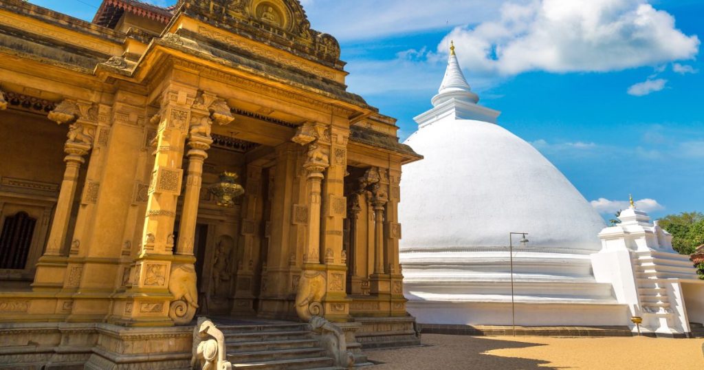 位於斯里蘭卡的 Kelaniya 廟宇，其金色柱廊細節精美，旁邊有一座潔白的圓形佛塔，天空晴朗，背景有藍天白雲，展示出佛教寺廟的莊嚴與典雅。