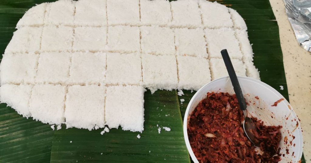 這張照片展示了切成小方塊的Kiribath(椰漿飯),旁邊擺放了一碗伴菜,背景為傳統的蕉葉餐桌佈置。