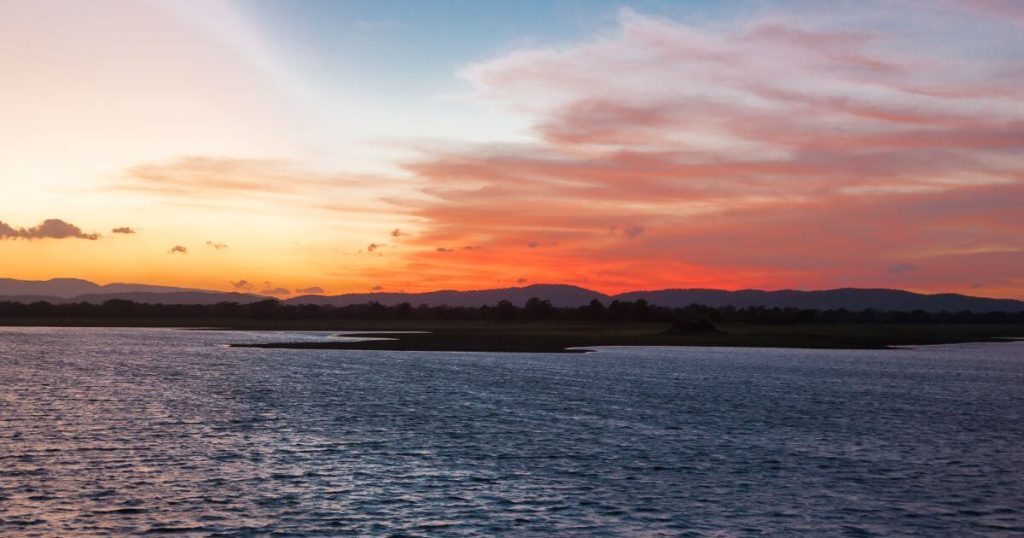 此圖片展示了斯里蘭卡帕拉卡拉馬薩穆德拉(Parakrama Samudra)湖面的壯麗日落景象,天空被染成橘紅色與藍色的漸層,與湖水的波光粼粼相映成趣。遠方的山脈和湖岸上的樹林增添了畫面的深度,呈現出一幅平靜而美麗的自然景致。