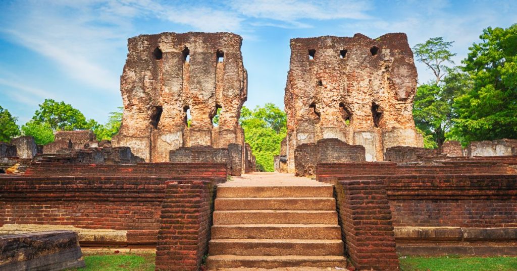 該圖片展示了斯里蘭卡帕拉克馬巴胡宮殿的壯麗遺跡。宮殿由紅磚建造而成，正中央的階梯通向殘破的宮牆，牆上依稀可見建築的雕刻細節。背景是青翠的樹林與湛藍的天空，整體氛圍充滿古老的歷史氣息，讓人不禁感受到當年王朝的輝煌。