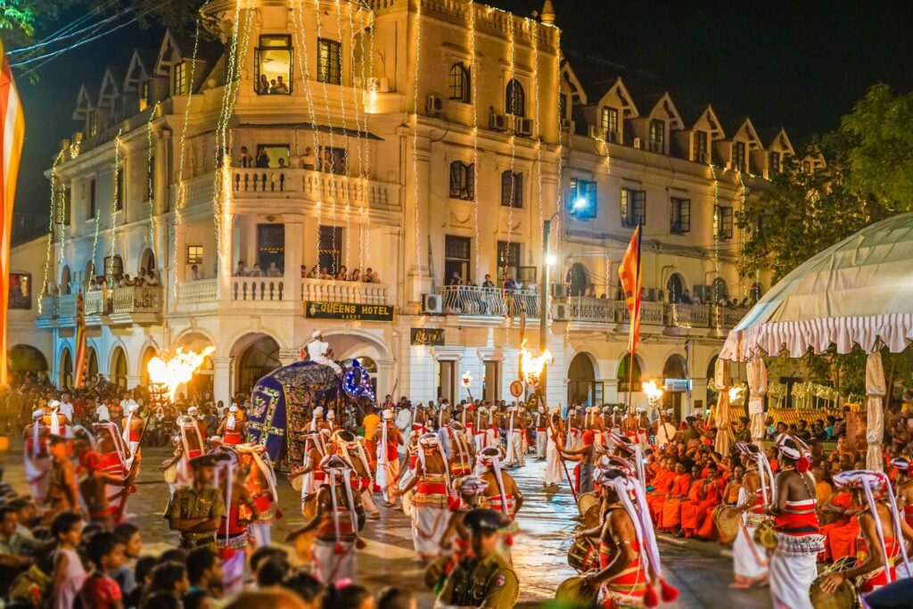 這張圖片展示了佛牙節中的遊行場景，舞者身著傳統服飾，隨著大象走過街道，旗幟在風中飄揚，沿途的觀眾也在欣賞這一場壯觀的慶典，這不僅是宗教活動，也是一場視覺和文化的饗宴。