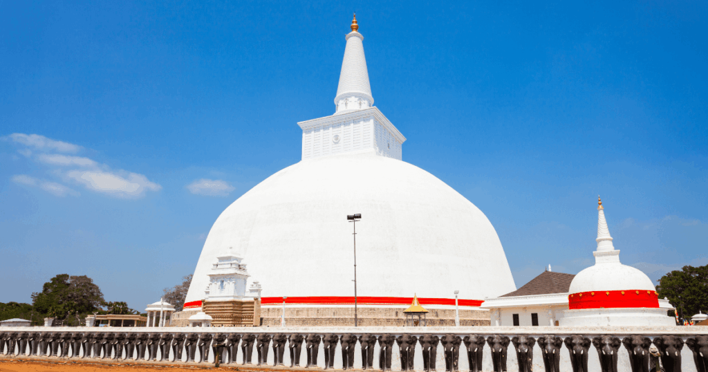 畫面中央是著名的魯梵維利沙雅大塔（Ruwanwelisaya），潔白的圓頂與高聳的尖塔在藍天下顯得格外莊嚴。塔身下方排列著數排大象雕像，周圍有佛教建築與樹木環繞，整體展現古都阿努拉德普勒的宗教與文化氛圍，這裡是斯里蘭卡重要的佛教朝聖聖地。