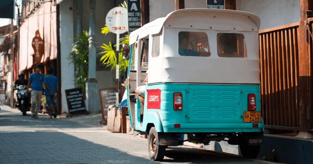 這張圖片展示了斯里蘭卡加勒(Galle)的街道,畫面中可以看到一輛典型的斯里蘭卡三輪車(嘟嘟車),其鮮明的藍綠色車身和街道的風景形成了鮮明的對比。街道兩側有當地的商店和居民,整個畫面充滿了熱帶風情,展現了加勒這座古城的日常景象。