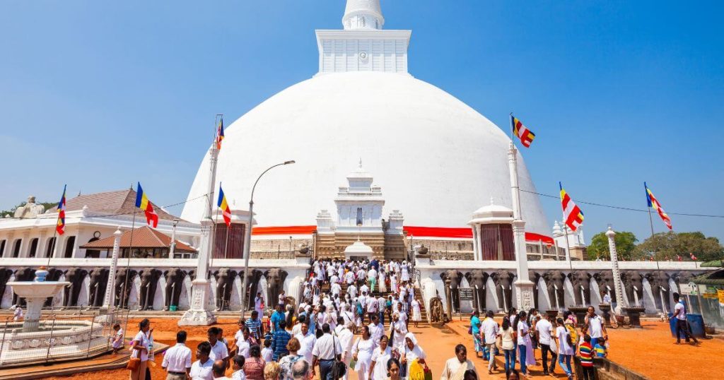 這張圖片展示了斯里蘭卡阿努拉德普勒著名的大寶塔。塔前聚集了大量前來朝聖的信徒,他們穿著白色衣服,象徵著虔誠與淨化。背景中,大寶塔潔白耀眼,四周懸掛著鮮豔的佛教旗幟,增添了這個重要宗教場所的神聖氛圍。