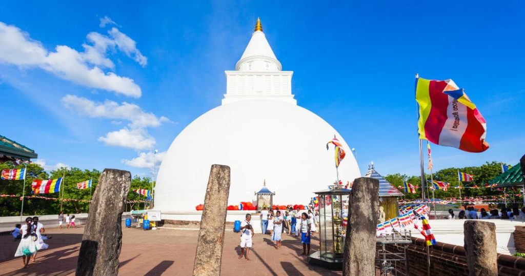 這張圖片展示了斯里蘭卡著名的卡塔拉伽馬佛塔,在藍天白雲的襯托下,白色的佛塔顯得格外壯麗。佛塔周圍飄揚著佛教旗幟,還有一些信徒在附近參拜,展現了當地濃厚的宗教氛圍。