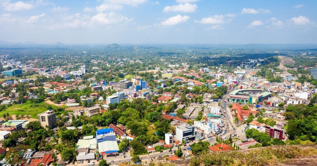 此圖片展示了斯里蘭卡庫魯內格勒市的全景，俯瞰整個城市，能看到市中心的各式建築及廣大的綠地。藍天白雲之下，遠處的山巒和河流隱約可見，反映出這個城市與自然環境的融合與和諧。