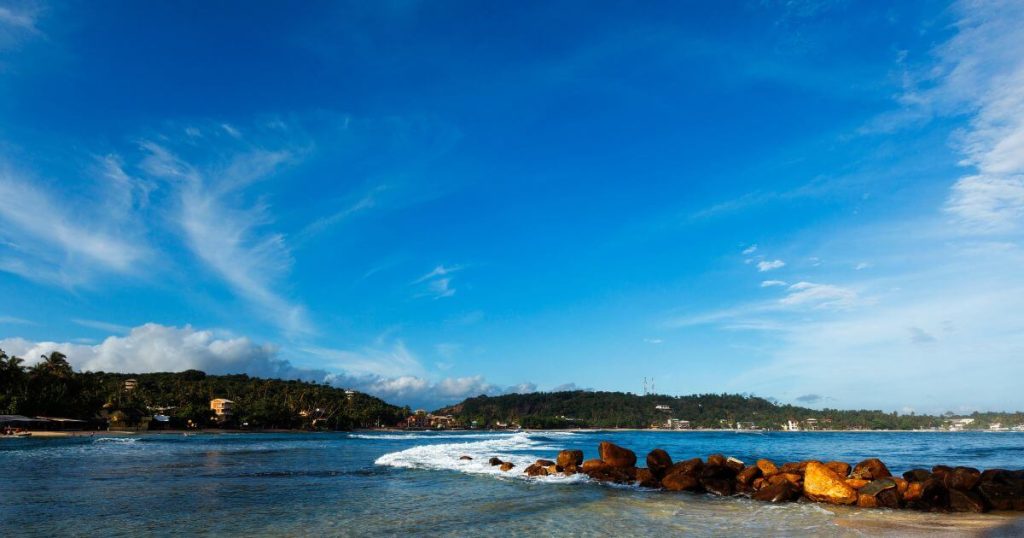 這張圖片展示了斯里蘭卡米瑞莎海灘的風景,天空碧藍,海浪輕輕拍打沙灘,周圍環繞著翠綠的樹木和山丘,展現了寧靜的海邊度假勝地。