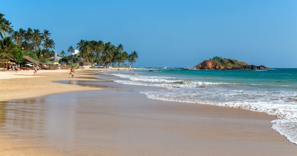 該圖片捕捉了斯里蘭卡美蕊沙海灘的平靜景象,潔白的沙灘和碧藍的海水完美融合。背景中搖曳的椰子樹與散步的遊客,勾勒出一幅悠閒的度假畫面,遠處的小島為這片海岸增添了一抹神秘的色彩。