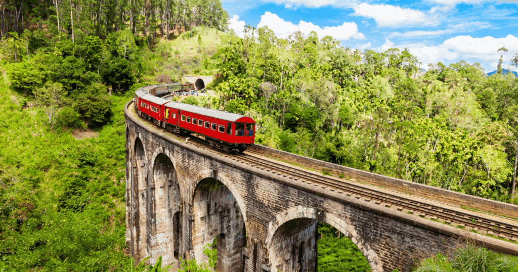 這張照片拍攝於斯里蘭卡(Sri Lanka)著名的九拱橋(Nine Arch Bridge),畫面中的紅色列車正行駛於古老的石橋之上,橋身拱形優美,位於綠意盎然的叢林與山林環繞中。天氣晴朗,藍天白雲搭配鮮紅列車構成強烈對比,展現出斯里蘭卡經典的慢旅風情與高原鐵道魅力。