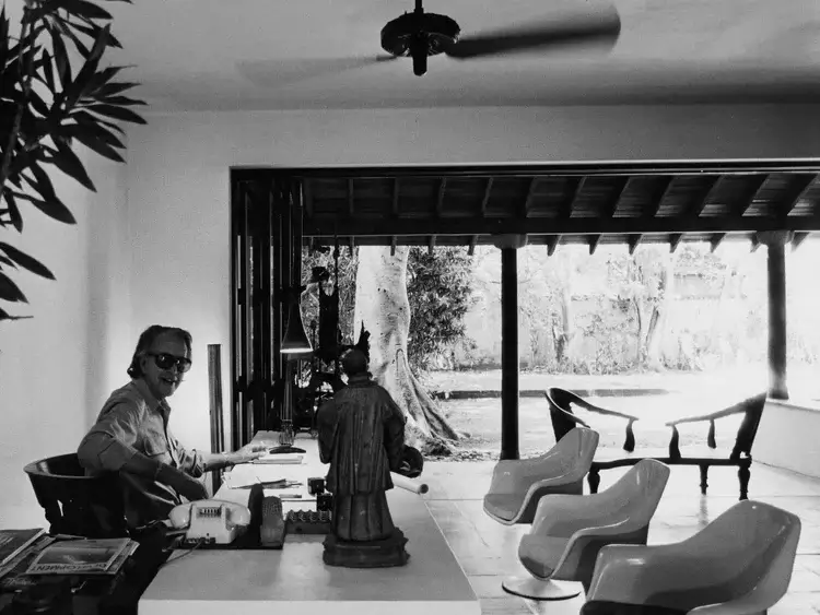 黑白照片中,斯里蘭卡建築師Geoffrey Bawa坐在書桌前,身後是半開放的庭院空間,展現他「熱帶現代主義」的設計理念。簡約的室內擺設、傳統木椅與現代塑料椅並置,呈現跨越時代的美感。桌上擺放佛像與書籍,與窗外自然景觀相互呼應,體現Bawa將自然、文化與建築融合的特色。這裡是他孕育靈感與設計的重要場所,也是理解其建築哲學的關鍵場景。