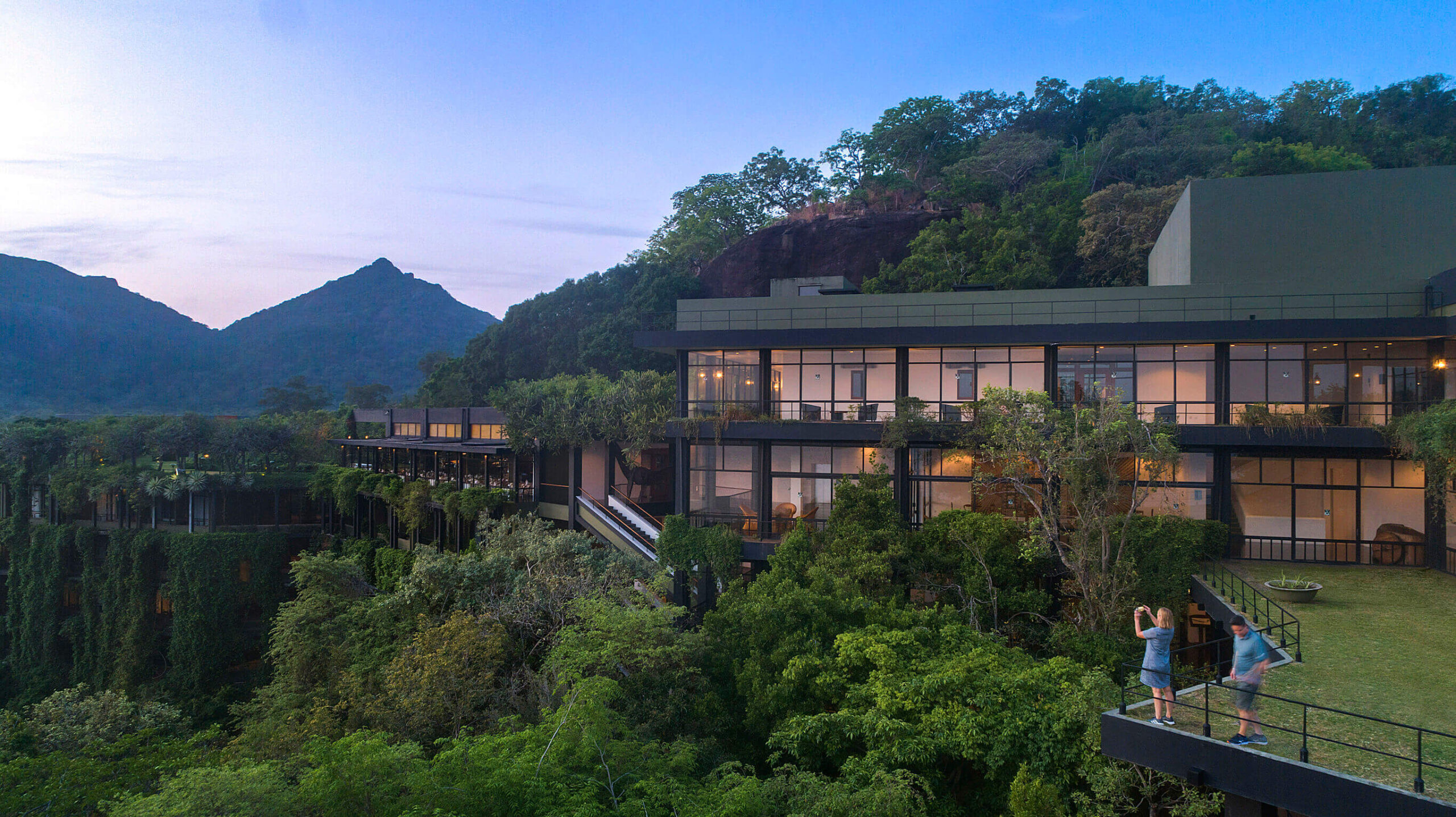 斯里蘭卡坎達拉馬酒店依山而建,外牆覆滿綠植,與周圍叢林完美融合。大片落地玻璃窗映照著山巒景色與夕陽餘暉,展現自然與建築的和諧之美。前方陽台上可見旅客欣賞風景、拍照留念,氛圍寧靜悠閒。
