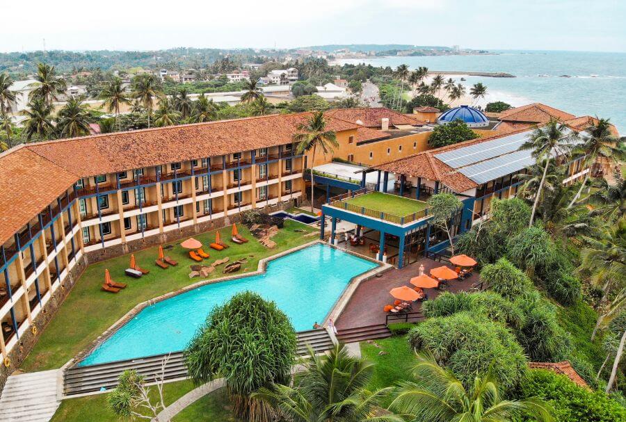 俯瞰斯里蘭卡傑特威燈塔，紅瓦屋頂與現代設計相融合，環抱著寬敞的藍色泳池，周圍佈滿熱帶棕櫚樹與綠意盎然的花園。遠方可見印度洋海岸線，營造出悠閒度假的氛圍。