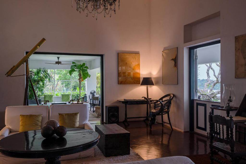 斯里蘭卡盧努甘卡莊園的室內空間充滿藝術氣息與古典韻味,白色牆面搭配深色木質家具,營造出溫馨又高雅的氛圍。房間內擺放黃綠抱枕的沙發、黑色圓桌以及黃銅色天文望遠鏡,展現主人的品味與生活格調。透過大面窗戶可欣賞綠意盎然的花園景色,讓室內外融為一體。