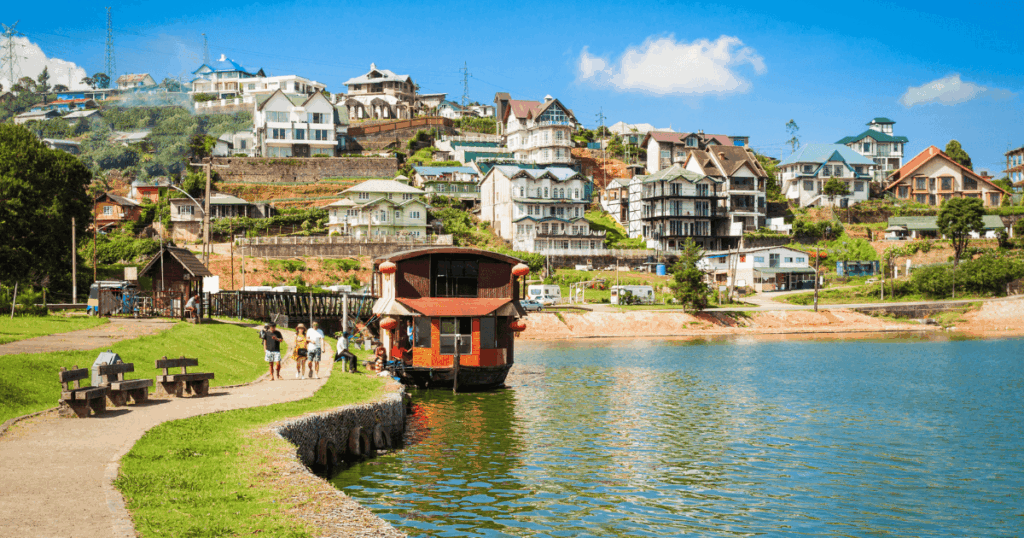 圖片展現斯里蘭卡努瓦勒埃利耶（Nuwara Eliya）湖畔的恬靜景色，湖水碧綠映照著藍天白雲，岸邊停泊著一艘紅色木船，吸引遊客駐足拍照。小鎮沿坡而建，整齊排列著帶有歐風建築特色的民宅，屋頂色彩多樣，綠意點綴其間。湖邊的步道與長椅增添休閒氛圍，整體呈現悠閒愜意的度假情境。