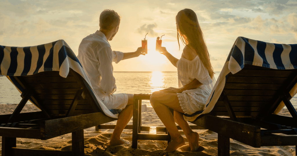 一對情侶坐在海邊的躺椅上，手中舉著果汁對飲，夕陽灑在海面上閃耀金光，氣氛浪漫而寧靜。畫面象徵蜜月旅行中專屬的親密時刻，展現兩人共享美好時光的幸福氛圍。