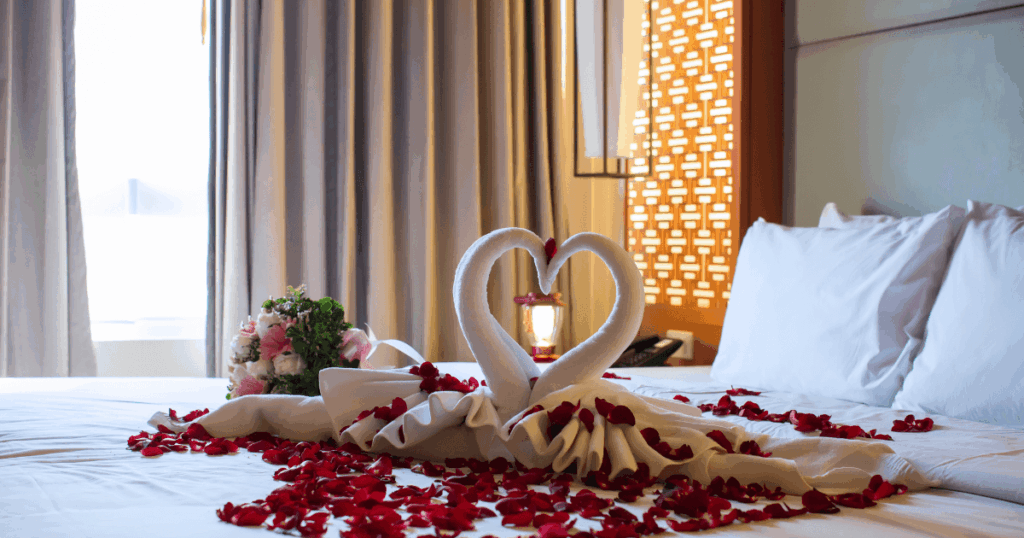 飯店房間內以白色毛巾摺成天鵝造型,形成愛心圖案,搭配滿床的玫瑰花瓣與鮮花花束,營造浪漫溫馨的蜜月氛圍,象徵幸福與愛情的開始。